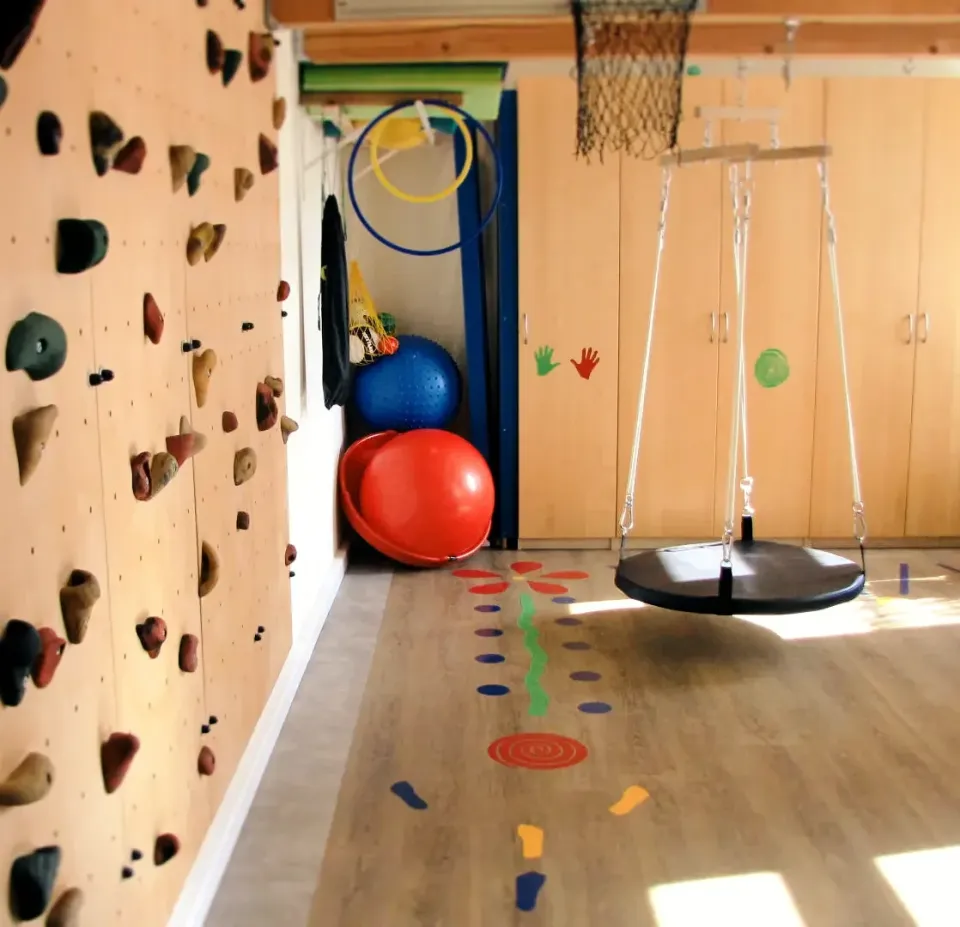 Therapieraum der Praxis mit Kletterwand, Schaukel, Bällen und bunten Bodenmarkierungen